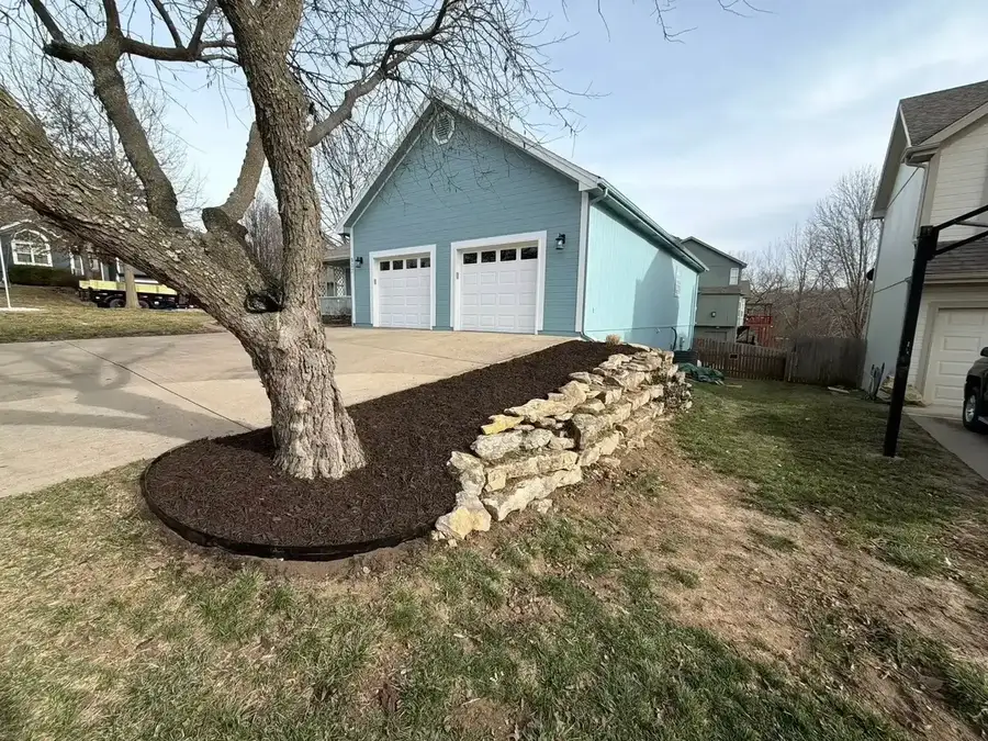 Natural fieldstone retaining wall with mulch bed and tree ring along driveway — retaining wall builder in Smithville MO